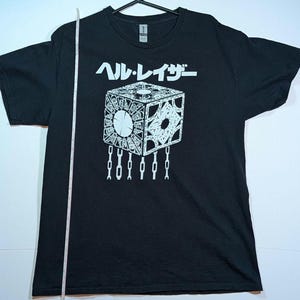 May include: Black t-shirt featuring a white graphic of a puzzle cube with chains and Japanese text. The shirt is a crew neck style and is hanging on a black hanger.