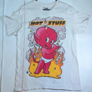 May include: White t-shirt featuring a cartoon devil character with red skin, horns, and a diaper. The shirt has the words "HOT STUFF" above the devil, with flames and stars in the design. The shirt is hanging on a black hanger.