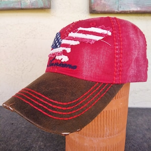 May include: Red baseball cap with a distressed American flag design and the word "Americana" embroidered on the front. The cap has a brown leather bill with red stitching.