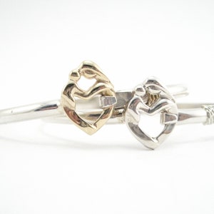 May include: Two silver bracelets with a heart-shaped design featuring a mother and child figure. One bracelet has a gold heart-shaped design, while the other is silver.