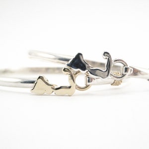 May include: A silver bangle bracelet with a gold state outline charm. The bracelet is twisted and has a small anchor charm.