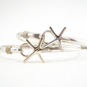 May include: Two silver-toned bangles with starfish accents. One bangle features a gold-toned accent near the clasp, while the other has a silver-toned accent. The starfish are the focal point, adding a nautical touch to the jewelry.
