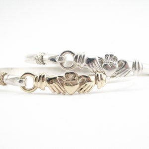 May include: Two silver Claddagh bracelets with a gold heart and hands on one bracelet. The bracelets are intertwined.