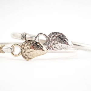 May include: Two silver bracelets with a gold and silver oyster charm. The bracelets are both thin and have a braided design.