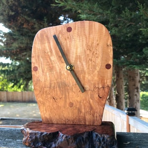 May include: A wooden clock with a unique, organic shape. The clock face is made of light wood with dark brown accents and a single black hand. The clock is mounted on a dark brown wooden base.