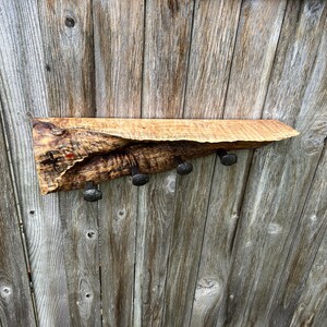May include: A rustic wooden shelf with four dark metal hooks. The shelf is made from a single piece of light brown wood with a natural edge. The hooks are evenly spaced along the bottom.