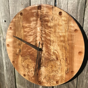May include: A round wooden clock with a black face and black hands. The clock face has a unique wood grain pattern with a light brown and dark brown color scheme. The clock is surrounded by a black rim and has a black border around the clock face.