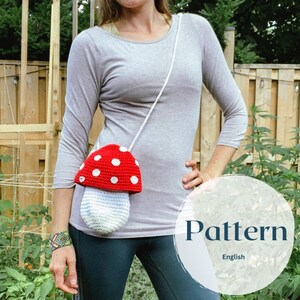 May include: A crocheted mushroom-shaped bag with a white strap. The bag is red with white polka dots and has a white stem. The bag is being worn by a person as a crossbody bag.