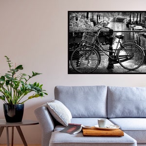 May include: A grey sectional sofa with a chaise lounge and a wooden tray on the chaise. A book and a cup of coffee are on the tray. A potted plant and a small table are in the foreground. A black and white photo of a bicycle on a bridge is hanging on the wall.