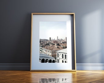 May include: A framed photograph of a cityscape with a view of buildings and a church steeple in the distance. The photo is in a light wood frame and is hanging on a gray wall.