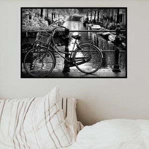 May include: A black and white photograph of a bicycle with a basket parked on a bridge over a canal. The bicycle is leaning against a railing and there are boats in the water.