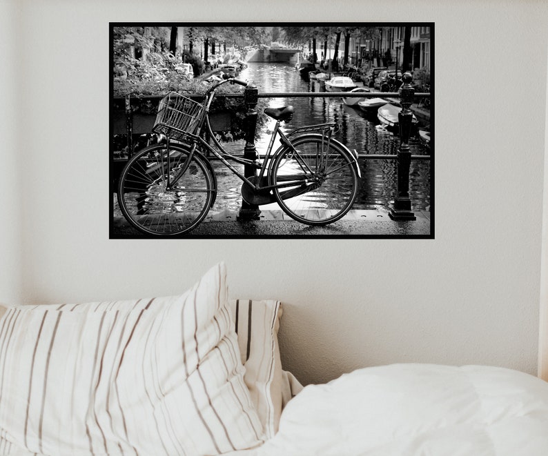 May include: A black and white photograph of a bicycle with a basket parked on a bridge over a canal. The bicycle is leaning against a railing and there are boats in the water.