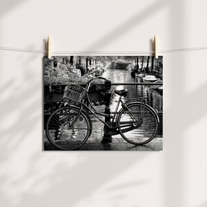 May include: A black and white photograph of a bicycle parked on a bridge over a canal. The bicycle has a basket on the handlebars and is leaning against a railing. The canal is lined with trees and buildings.