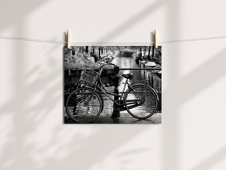 May include: A black and white photograph of a bicycle parked on a bridge over a canal. The bicycle has a basket on the handlebars and is leaning against a railing. The canal is lined with trees and buildings.
