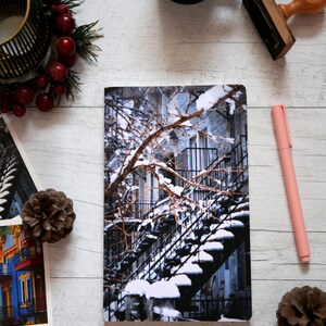 A winter notebook featuring a black and white photograph of Montreal's snow-covered spiral walk-up stairs with parked bicycles covered in snow. The notebook is surrounded by other Montreal notebooks and mini prints that can be framed or sent as cards.

The hand-bound notebooks measure 5x8 inches. They contain 48 pages of cream-white acid free paper with a smooth finish. The matte cover is smooth and scuff-resistant!

Each notebook comes with an insert containing 15 insightful journaling prompts!
