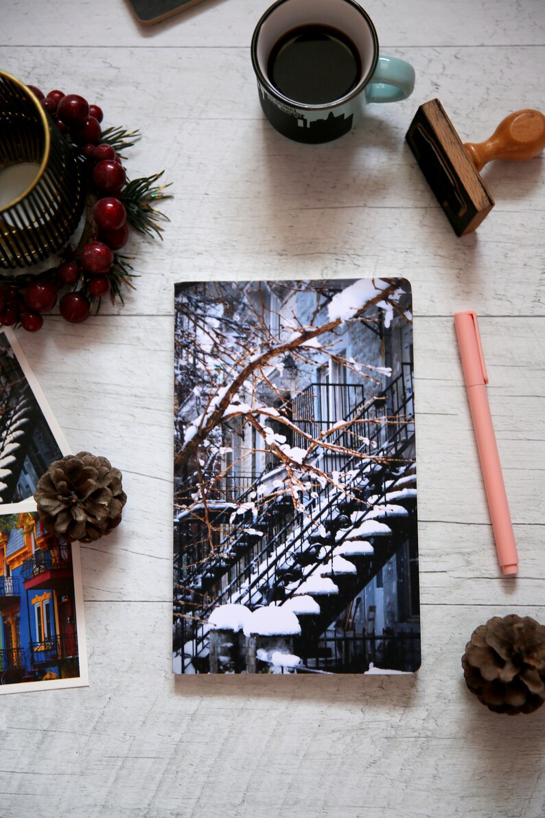 A winter notebook featuring a black and white photograph of Montreal's snow-covered spiral walk-up stairs with parked bicycles covered in snow. The notebook is surrounded by other Montreal notebooks and mini prints that can be framed or sent as cards.

The hand-bound notebooks measure 5x8 inches. They contain 48 pages of cream-white acid free paper with a smooth finish. The matte cover is smooth and scuff-resistant!

Each notebook comes with an insert containing 15 insightful journaling prompts!