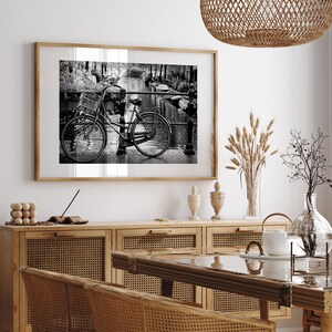 May include: Black and white photograph of a bicycle with a basket on the handlebars, parked on a cobbled street next to a canal. The photo is framed in a wooden frame and hanging on a white wall.