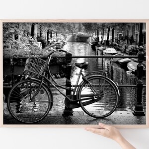 May include: A black and white photograph of a bicycle parked on a bridge over a canal in Amsterdam. The bicycle has a basket on the front and is leaning against a railing. The canal is lined with trees and buildings.