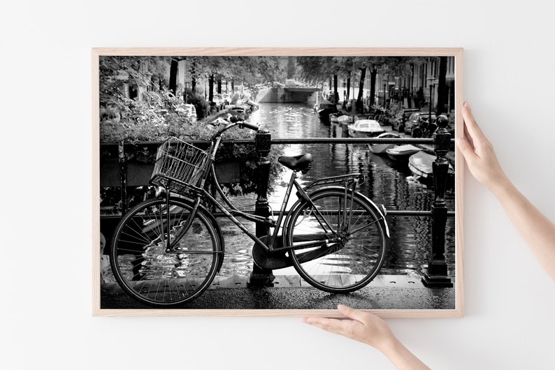 May include: A black and white photograph of a bicycle parked on a bridge over a canal in Amsterdam. The bicycle has a basket on the front and is leaning against a railing. The canal is lined with trees and buildings.