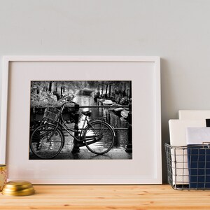 May include: Black and white photograph of a bicycle parked on a bridge over a canal. The bicycle has a basket on the front and is leaning against a railing. The canal is lined with trees and buildings.