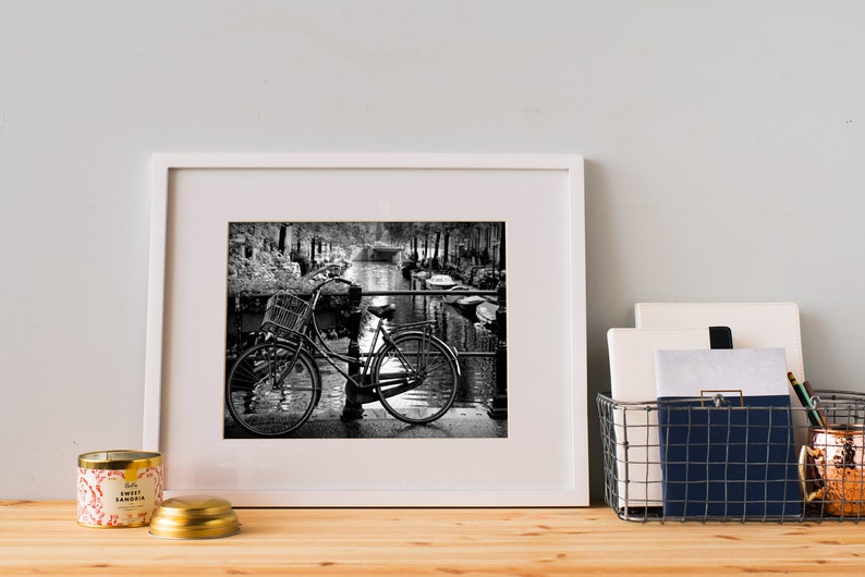 May include: Black and white photograph of a bicycle parked on a bridge over a canal. The bicycle has a basket on the front and is leaning against a railing. The canal is lined with trees and buildings.