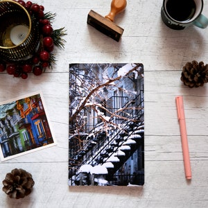 A winter notebook featuring a black and white photograph of Montreal's snow-covered spiral walk-up stairs with parked bicycles covered in snow. The notebook is surrounded by other Montreal notebooks and mini prints that can be framed or sent as cards.

The hand-bound notebooks measure 5x8 inches. They contain 48 pages of cream-white acid free paper with a smooth finish. The matte cover is smooth and scuff-resistant!

Each notebook comes with an insert containing 15 insightful journaling prompts!