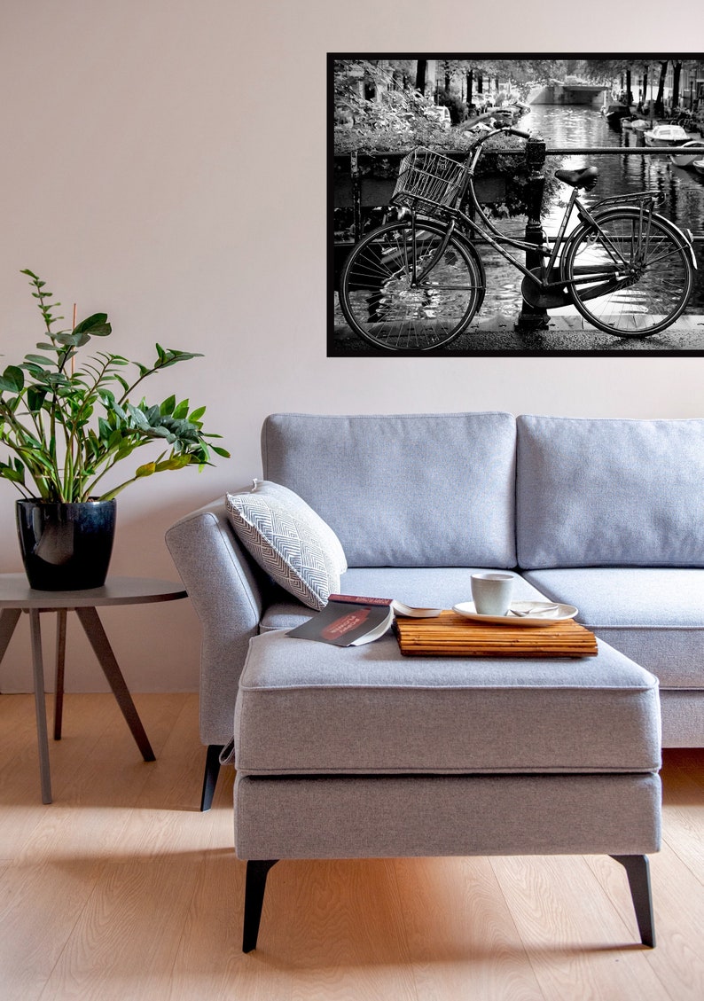 May include: A grey sectional sofa with a chaise lounge and a wooden tray on the chaise. A book and a cup of coffee are on the tray. A potted plant and a small table are in the foreground. A black and white photo of a bicycle on a bridge is hanging on the wall.