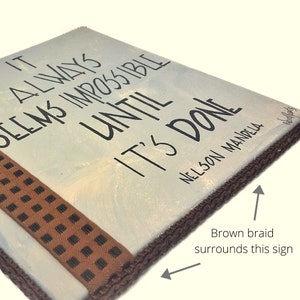 May include: A white canvas sign with the quote "It always seems impossible until it's done - Nelson Mandela" in black lettering. The sign is surrounded by a brown braid.