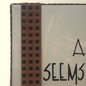 May include: A white canvas sign with brown trim and a black and brown patterned ribbon. The sign reads "It always seems impossible until it's done."