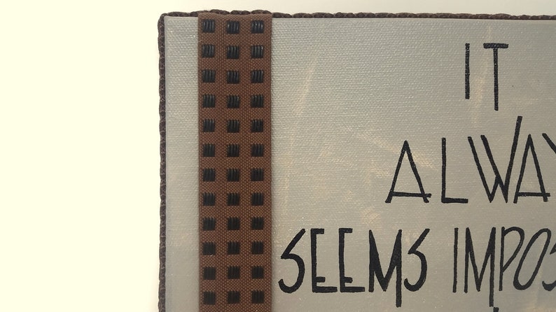May include: A white canvas sign with brown trim and a black and brown patterned ribbon. The sign reads "It always seems impossible until it's done."