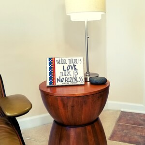May include: A wooden sign with a colorful border and the text "Where there is love there is no darkness. African proverb" on a wooden end table with a stone on top.