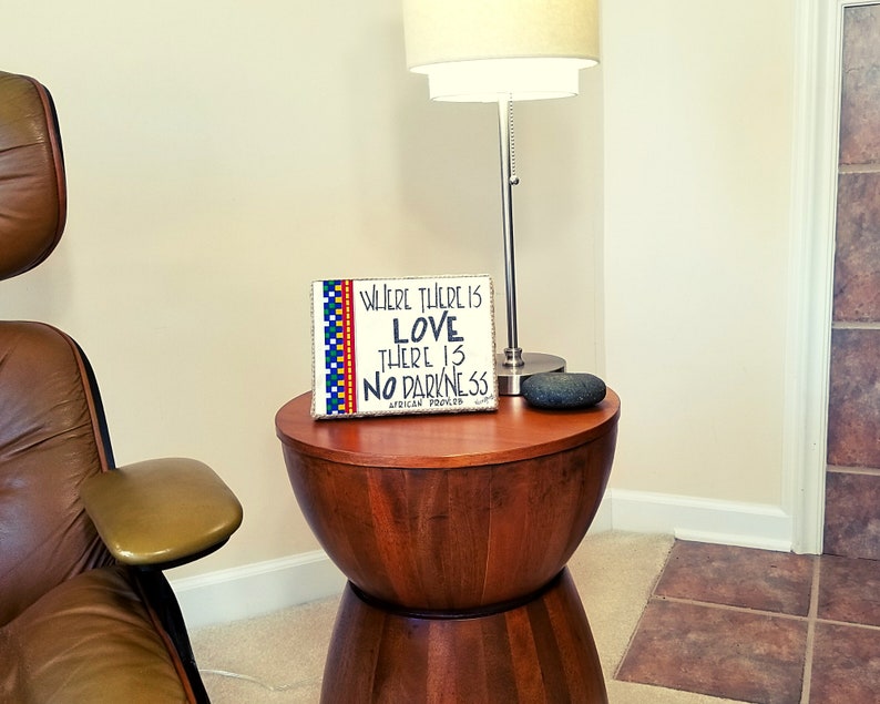 May include: A wooden sign with a colorful border and the text "Where there is love there is no darkness. African proverb" on a wooden end table with a stone on top.