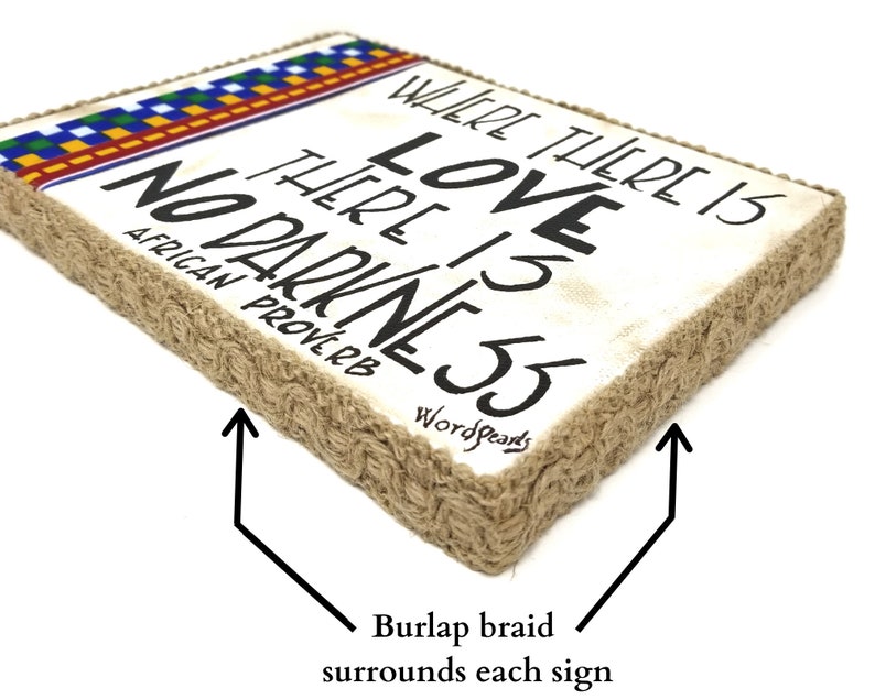 May include: A white wooden sign with the text "Where there is love there is no darkness" in black lettering. The sign is surrounded by a brown burlap braid and has a colorful patterned border at the top. The text "African Proverb" is written below the main text and "Wordsparks" is written below that.