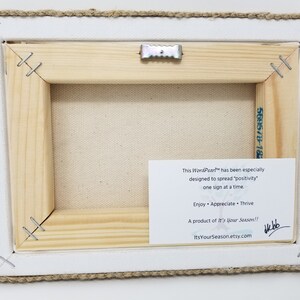 May include: A wooden framed canvas print with a white background and a burlap border. The back of the canvas has a white card with the text "This Won'Port&trade; has been especially designed to spread "positivity" one sign at a time. Enjoy &bull; Appreciate &bull; Thrive A product of It's Your Season!! ItsYourSeason.etsy.com" and a handwritten signature.