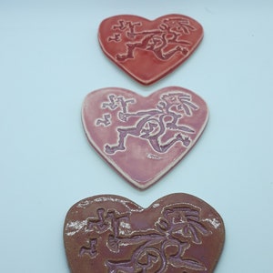 Puede incluir: Tres adornos de cerámica en forma de corazón con un esmalte rosa, rojo y marrón. Cada corazón presenta un diseño estilizado de una figura corriendo.