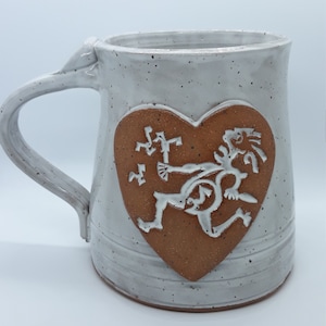 May include: White ceramic mug with a brown heart-shaped design featuring a stylized figure in white. The figure is running and has a heart-shaped head.