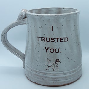 Puede incluir: Taza de cerámica blanca con un esmalte moteado. La taza tiene un asa y dice "I TRUSTED YOU." en letras negras. Hay una pequeña ilustración en blanco y negro en la parte inferior de la taza.