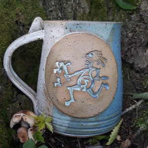 Puede incluir: Una taza de cerámica azul y blanca con un círculo marrón en el lateral que presenta una figura estilizada. La taza tiene un esmalte moteado y un asa.