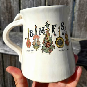 May include: White ceramic mug with a colorful graphic of musical instruments and flowers. The text "B.M.F.F.S" is printed on the mug. The text "FEBRUARY 2023" and "ASHEVILLE, NC" are also printed on the mug.