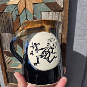 Puede incluir: Una taza de cerámica con un esmalte azul oscuro y marrón. La taza presenta un diseño circular blanco con una ilustración negra de una figura corriendo. El asa es azul. La taza se sostiene en una mano.