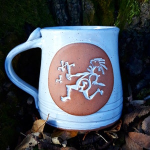 Puede incluir: Taza de cerámica blanca con un asa grande y un diseño circular marrón. El diseño presenta una figura blanca estilizada en movimiento. La taza tiene una textura moteada y un borde ancho.