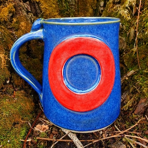 May include: A blue ceramic mug with a red circle design. The mug has a handle and is sitting on a bed of moss and twigs.