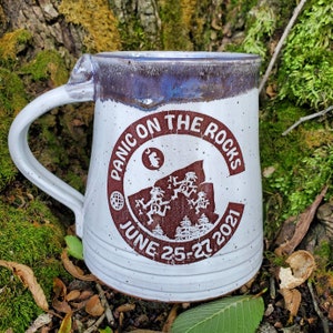 May include: White ceramic mug with a brown and white logo that says "Panic on the Rocks June 25-27 2021". The logo features a mountain range with stick figures dancing on top.