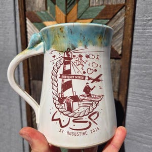Peut inclure: Une tasse en céramique blanche avec un glaçage bleu et vert. La tasse présente une illustration brune d'un phare, d'un bateau et le texte "She's Got Wings" et "WSP St. Augustine 2025".