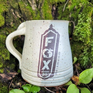 May include: A white ceramic mug with brown speckles and a brown and blue logo that says "FOX" with a small apple graphic and the text "www.810-813-22" below. The mug has a handle and is sitting on a bed of green moss.