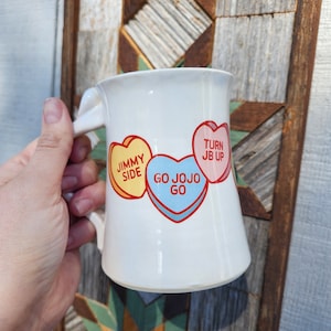 May include: White ceramic mug with a handle. The mug has three candy heart designs on it. The hearts say "Jimmy Side", "Go Jojo Go", and "Turn JB Up".