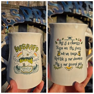 May include: Cream-colored ceramic mug with a curved handle. One side features the text "WSMFP" above a rainbow, flower, and mountain scene. The other side has the text "This is a cleaning..." with floral and butterfly illustrations.