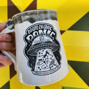 May include: White ceramic mug with a black and white illustration of a flying saucer with a person inside. The text "Doom Cosmic Panic" is written in a retro font with a planet symbol. The text "May 5-6 Charleston, SC 2023" is written below the illustration.
