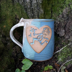 Puede incluir: Una taza de cerámica azul y marrón con un diseño en forma de corazón que presenta una figura estilizada. La taza tiene un asa y un esmalte moteado.