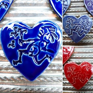 Puede incluir: Azulejos de cerámica en forma de corazón en tonos azules y rojos. Cada azulejo presenta un diseño estilizado en relieve. Los azulejos están dispuestos sobre una superficie metálica texturizada. El acabado brillante realza los colores vibrantes y los detalles intrincados.
